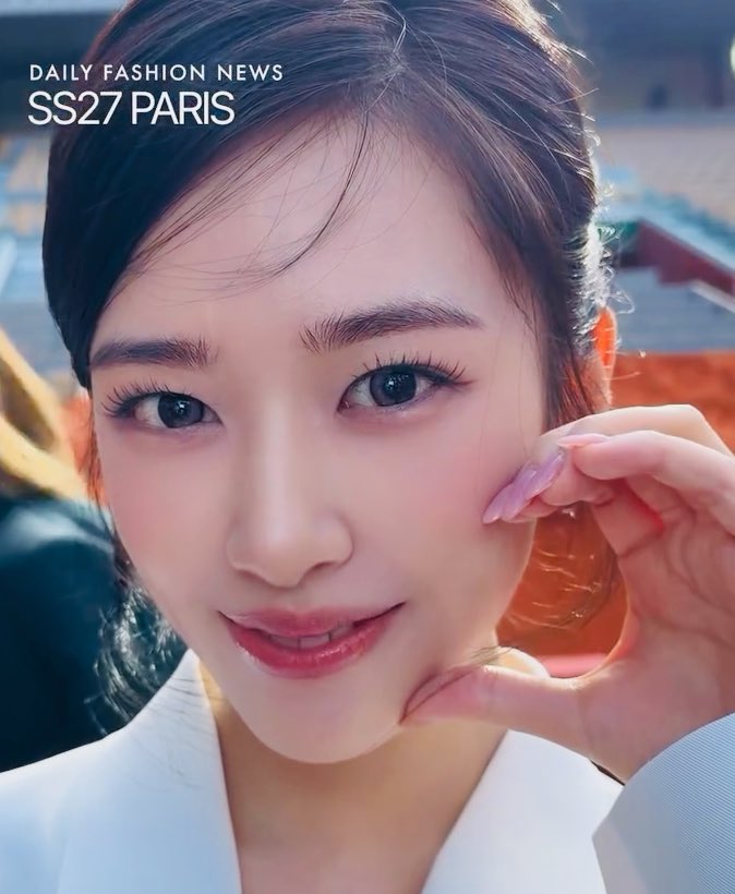A close-up selfie-style portrait of a young Asian woman with dark hair touching her face, with text reading 'DAILY FASHION NEWS SS27 PARIS' in the upper left corner.