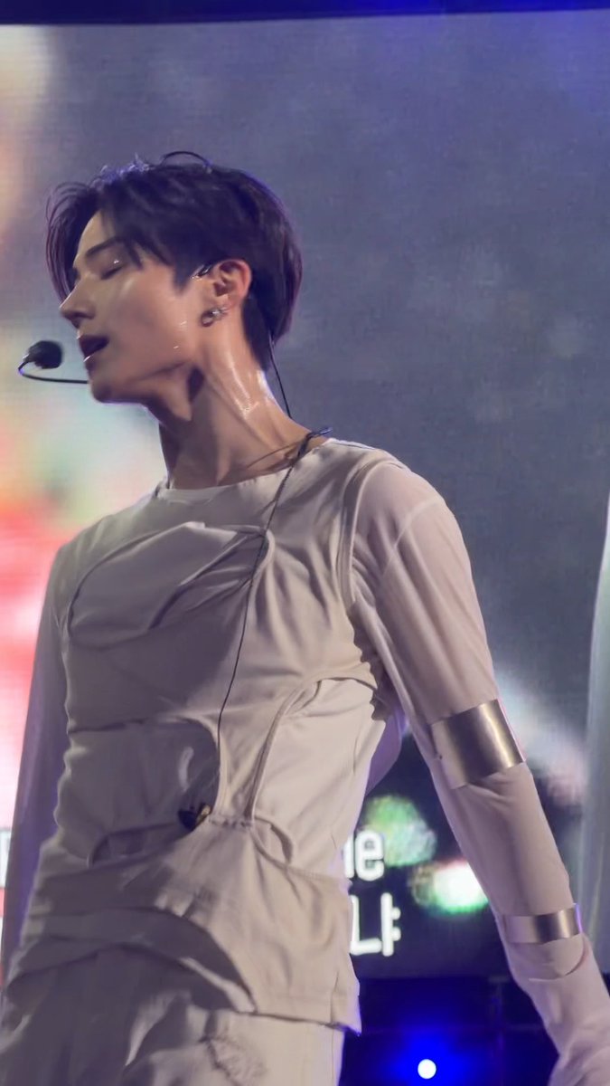 A K-pop performer on stage wearing a light grey/beige sheer outfit with metallic silver arm cuffs, dark lipstick, and a headset microphone, captured mid-performance with stage lighting in the background.