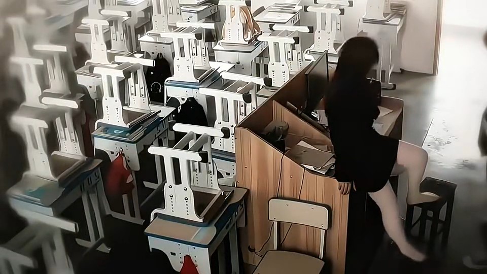 A dimly lit classroom is filled with stacked desks and chairs, while a person in dark clothing sits on a stool in the foreground.