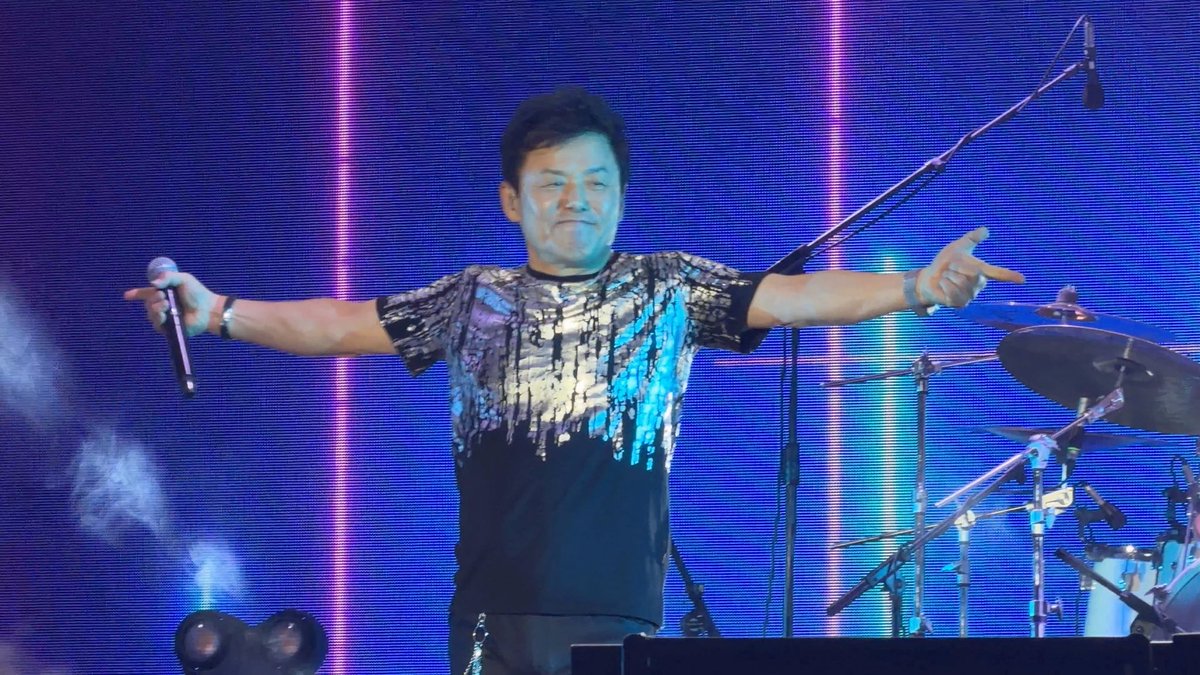 A male singer in a sparkly sequined shirt stands on a stage with arms outstretched, holding a microphone, set against a blue illuminated background with a drum set visible behind him.