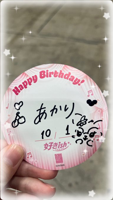 A hand holding a circular pink sticker that reads "Happy Birthday!" with the date "10/1" and handwritten Japanese text, along with the "AKB48" logo.