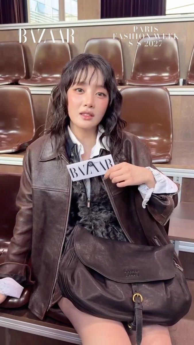A woman with wavy dark hair wearing a brown leather jacket holds a card with 'BAZAAR' text, with 'BAZAAR' and 'PARIS FASHION WEEK S/S 2027' displayed at the top, seated in brown stadium-style seats.