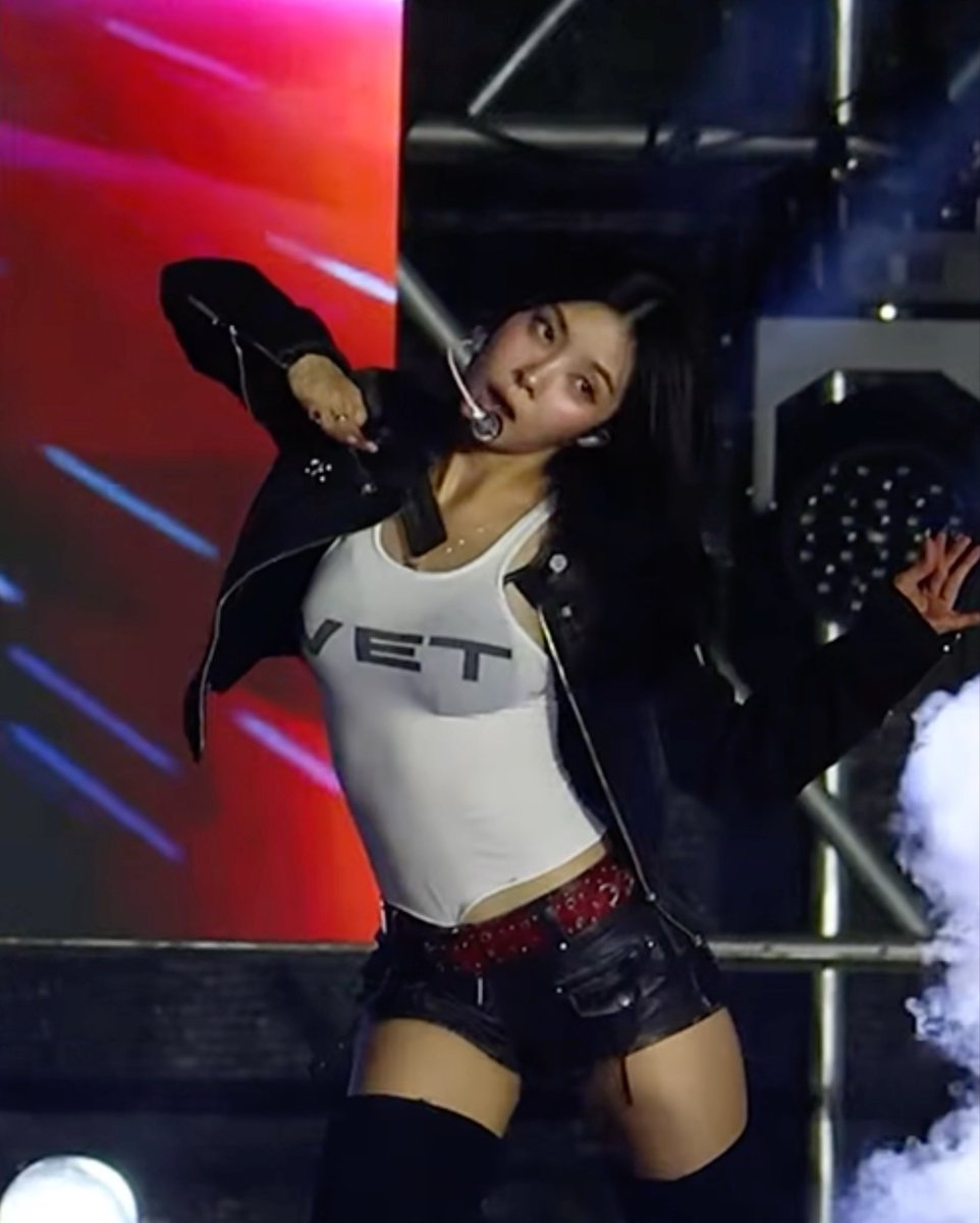 A K-pop performer on stage wearing a white tank top with 'VET' text, black jacket, and black shorts, dancing with a headset microphone.
