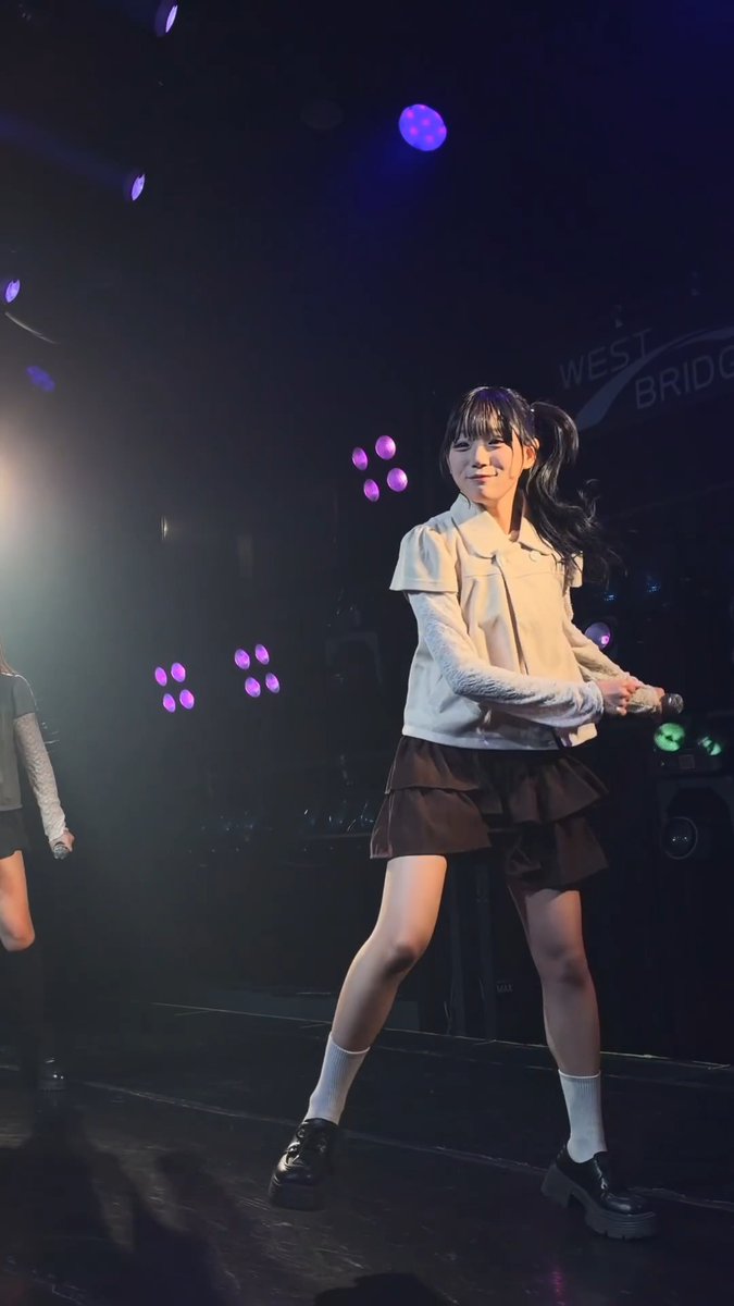 A young woman with dark pigtails wearing a white blouse, black skirt, white knee socks, and black shoes poses with a smile on a stage under colorful lights; 'WEST BRIDGE' is partially visible in the background.