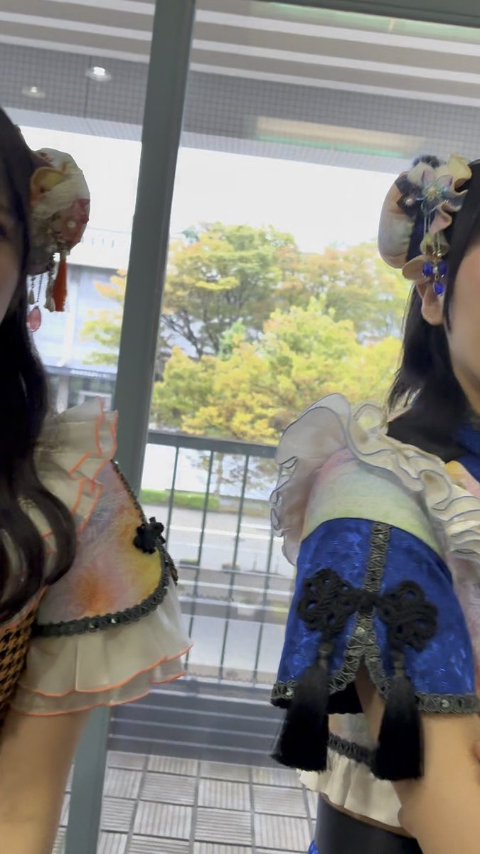 Two people wearing elaborate, colorful Lolita-style outfits with hair accessories and frilled collars stand near a large window overlooking a green outdoor area.