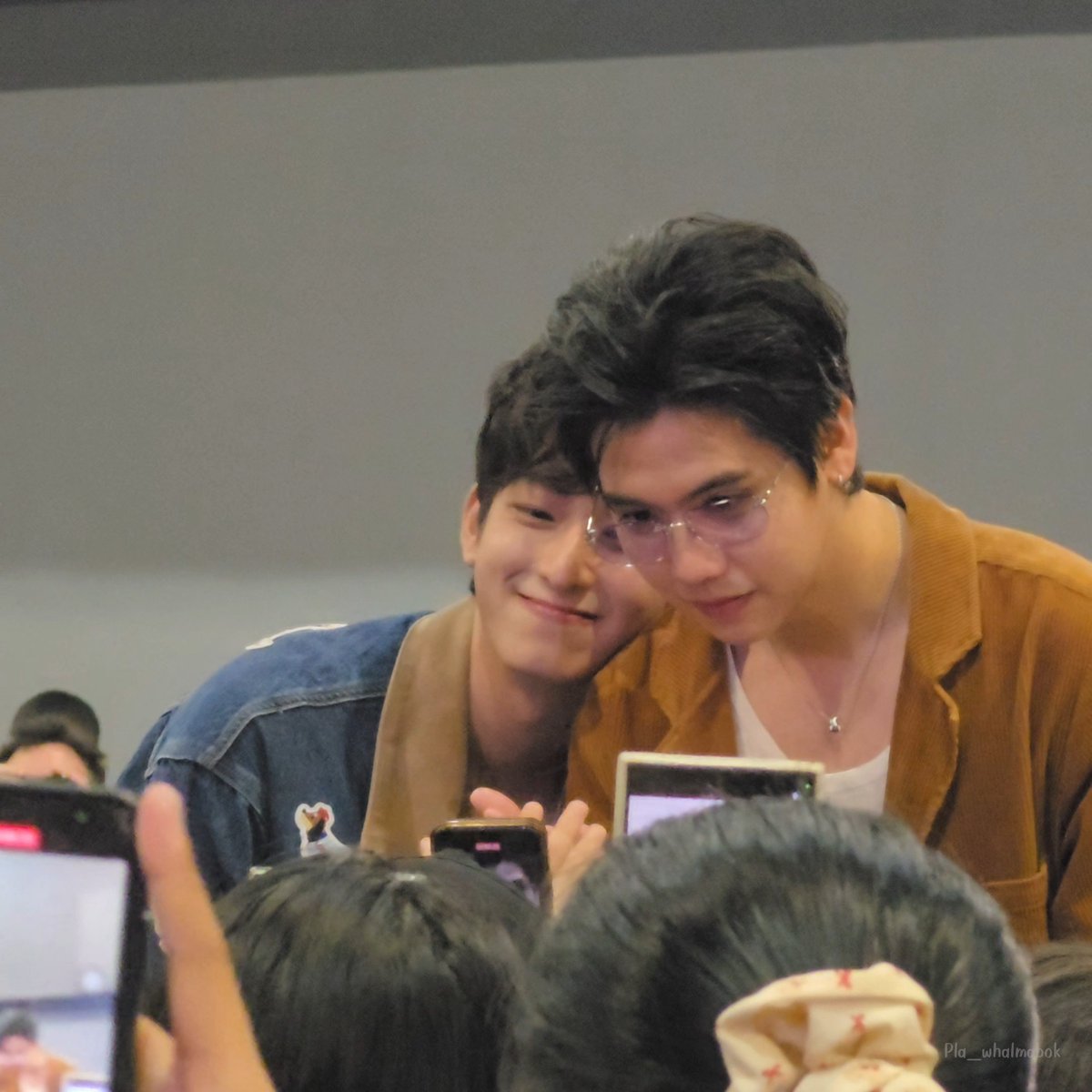 Two young men smiling while posing for photos with fans, one wearing a denim jacket and the other wearing a brown jacket with glasses and a necklace, with a watermark 'Pla_whalmabook' in the corner.