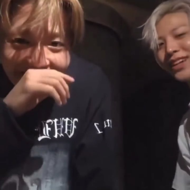 Two young men sit together in a dark setting; the one on the left wears a black sweatshirt with white Gothic-style text and covers his mouth with his hand, while the blonde-haired man on the right smiles.