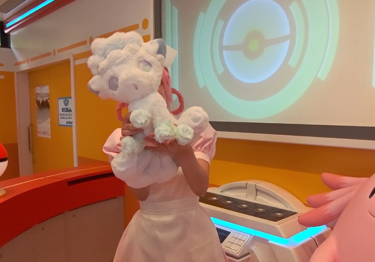 A person dressed in a pink Pokémon Nurse Joy costume holds up a white Vulpix Alolan plush toy in a themed room with a Pokéball emblem on the wall and a Chansey figure visible.