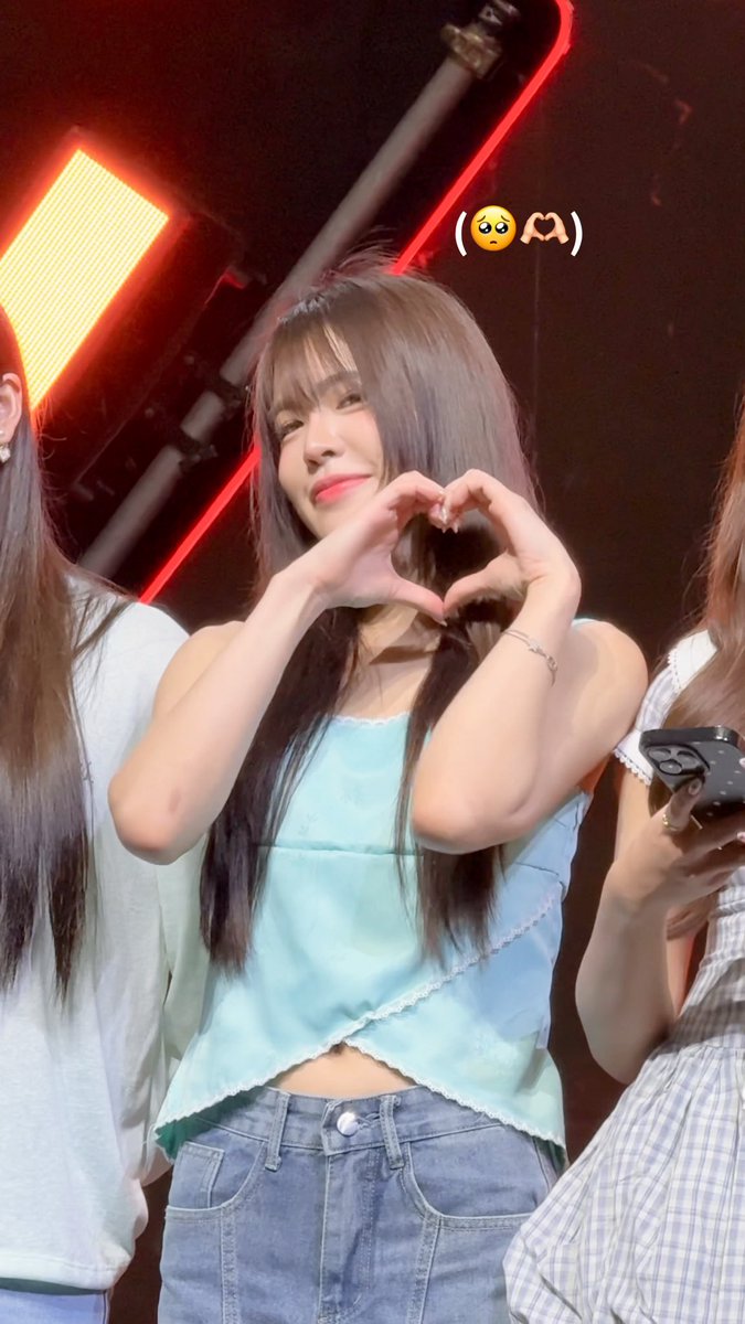 A young woman with long dark hair, identified by the hashtag as Arlee from BNK48, smiles brightly while making a heart gesture with her hands on a stage lit with red lighting. An emoji overlay "(🥺🫶)" appears near her head, matching the cute, affectionate vibe of the caption about being lovely with stars.