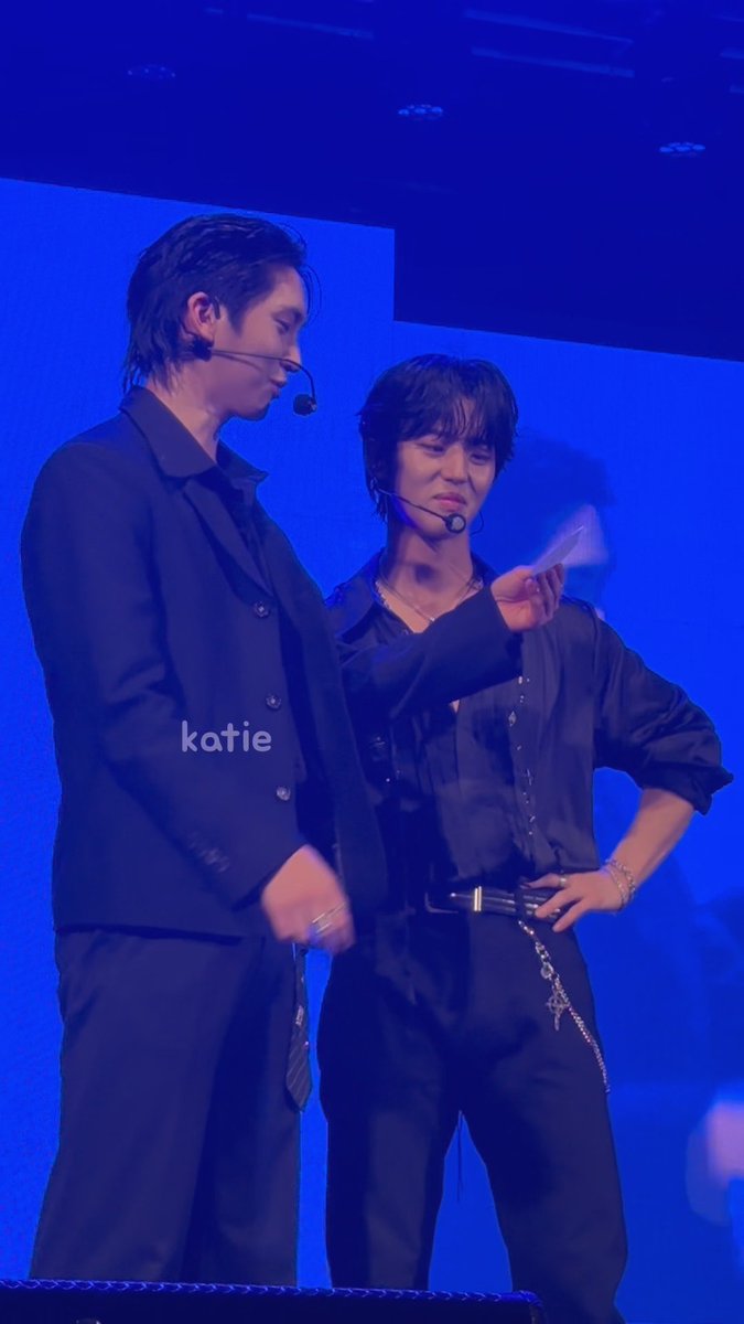 Two performers on stage wearing black outfits and headset mics; one points while speaking to the other, with the watermark 'katie' visible on the image.