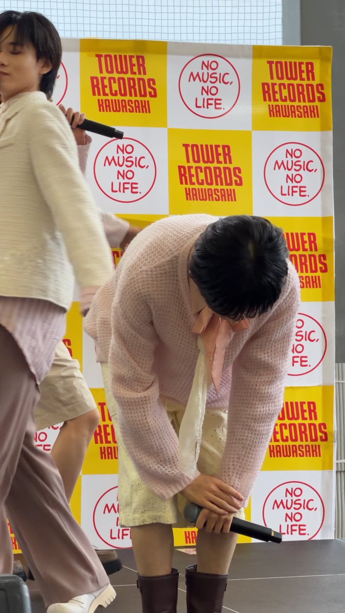 A performer in a pink sweater and patterned shorts bows deeply while holding a microphone, with another performer in cream clothing partially visible on the left, all set against a Tower Records Kawasaki 'NO MUSIC, NO LIFE' backdrop.