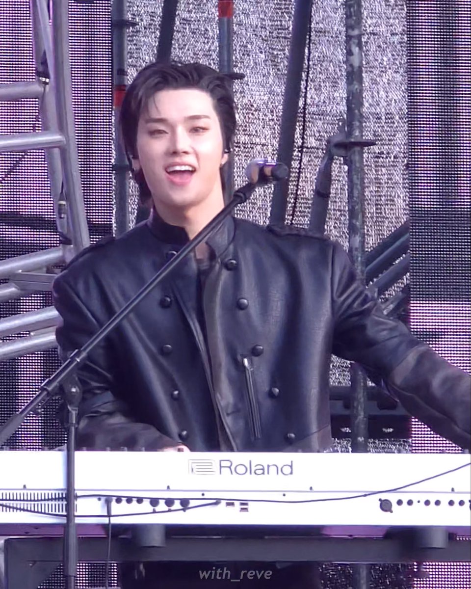 A young man with dark hair performing on stage behind a Roland keyboard, smiling while wearing a black leather jacket, with the watermark 'with_reve' visible.