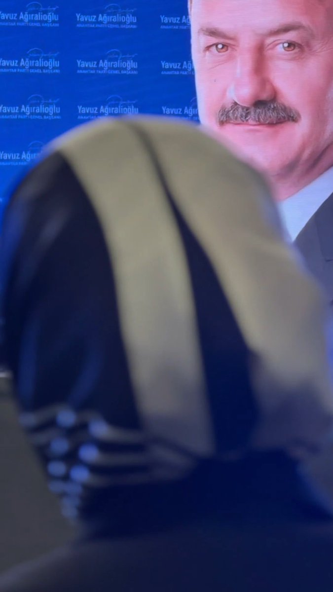 A televised broadcast showing the back of a woman wearing a black and yellow striped headscarf in the foreground, facing a large screen displaying a portrait of a man with a mustache against a backdrop repeatedly branded with "Yavuz Ağıralioğlu, SİVAS Milletvekili Genel Başkanı."