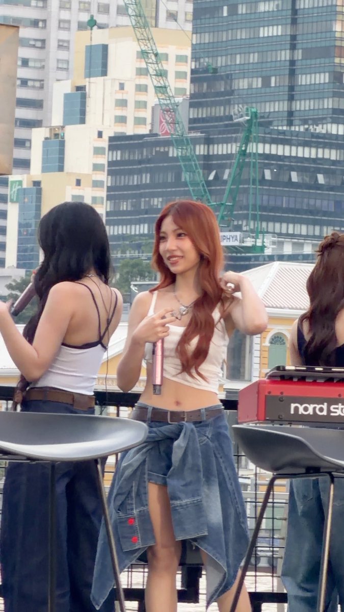 A woman with long reddish-brown hair smiles while holding a microphone on an outdoor rooftop, with a city skyline and construction cranes in the background, near a Nord Stage keyboard.