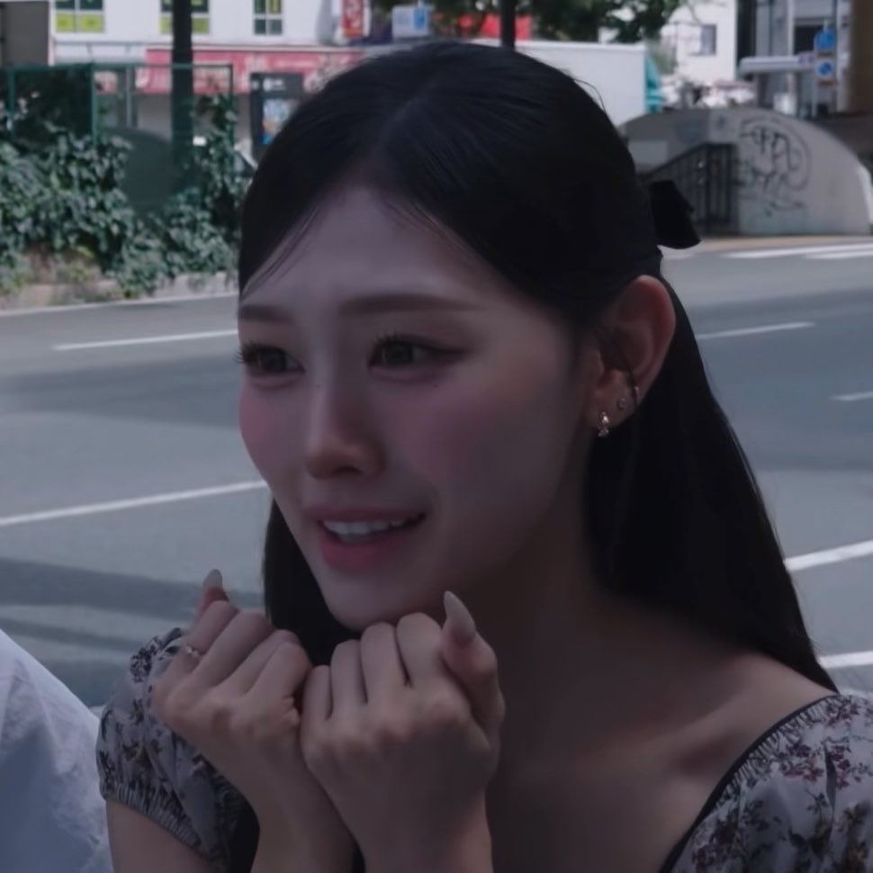 A young woman with dark hair and a black bow smiles emotionally on a city street, hands clasped near her chin.