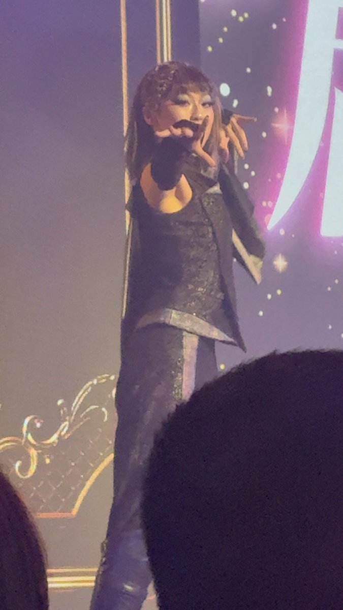 A female performer on stage wearing a dark outfit with silver accents, holding a microphone and posing dramatically with one hand raised, against a starry backdrop with audience members visible in the foreground.