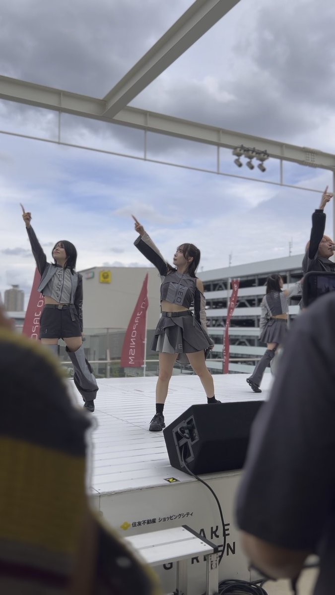 Three female performers in gray futuristic costumes perform on an outdoor stage, all pointing upward, with red flags reading 'MAISHOWEN' visible in the background.