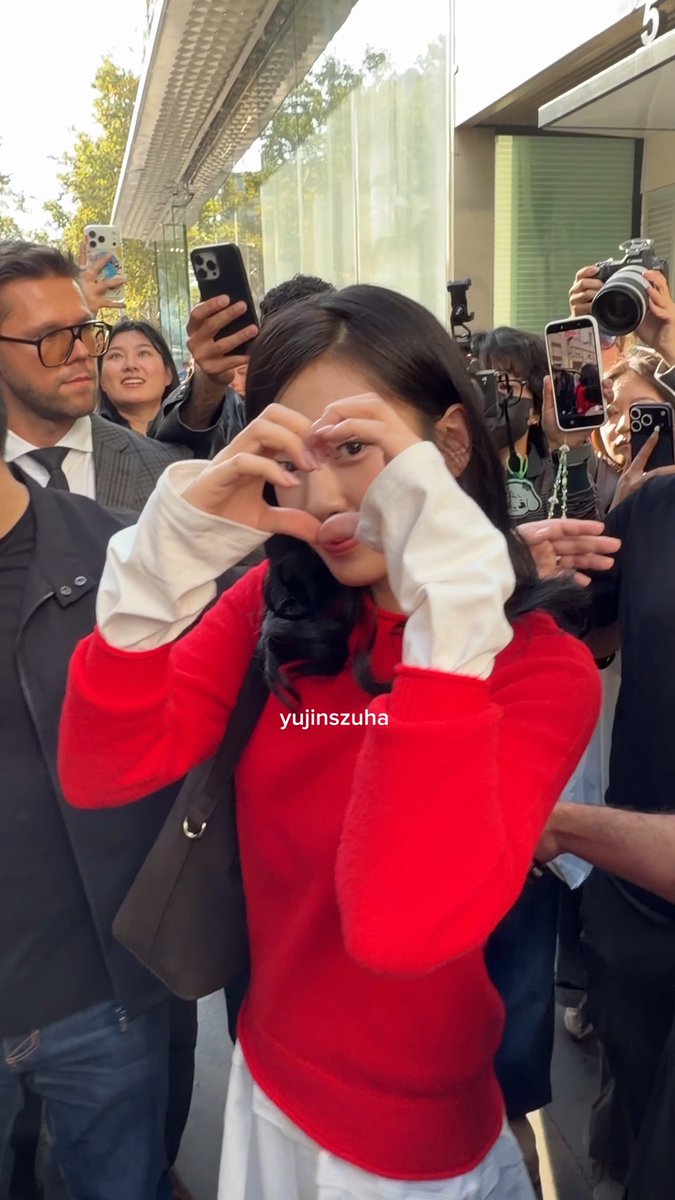 A young woman in a red sweater over a white collared shirt makes a heart shape with her hands in a crowd of people taking photos with smartphones, with the text 'yujinszuha' overlaid in the center.