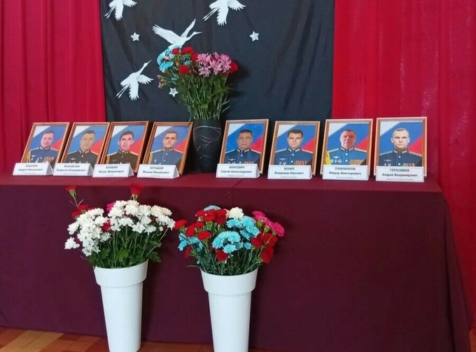 A memorial display features eight framed portraits of Russian military servicemen in uniform, arranged in a row on a maroon-draped table with nameplates beneath each photo, accompanied by bouquets of red, white, and pink flowers in tall vases and a decorative backdrop with white bird silhouettes, honoring the crew members killed in the recent Tu-95MS crash in Amur Oblast.