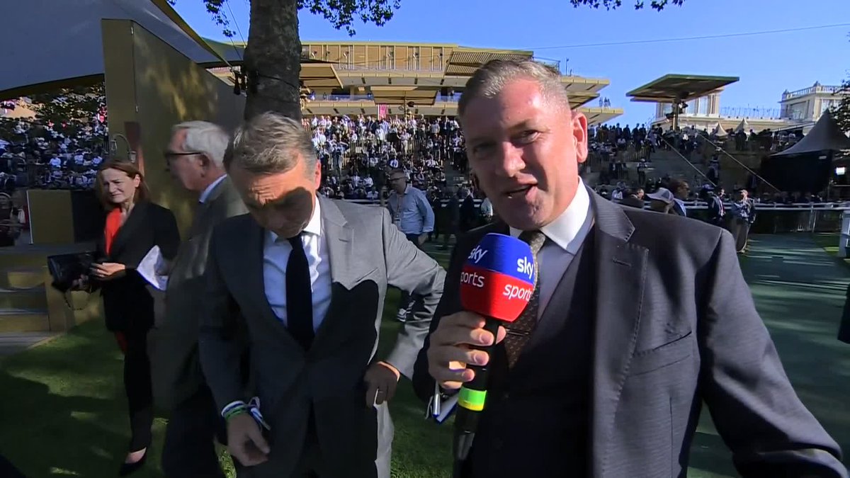 A Sky Sports reporter in a suit holds a microphone while another man in a grey suit looks down, set against a crowded outdoor venue resembling Royal Ascot.
