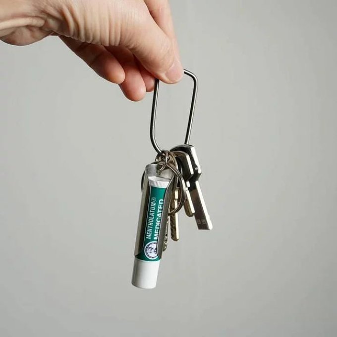 A hand holds a carabiner attached to a keychain featuring keys and a small tube of 'METHOGELATED' medicated lip balm.