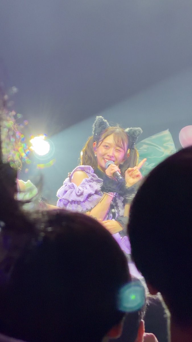 A female performer sings into a microphone on stage, wearing a purple and black outfit with cat-ear headbands, while audience members' silhouettes are visible in the foreground.