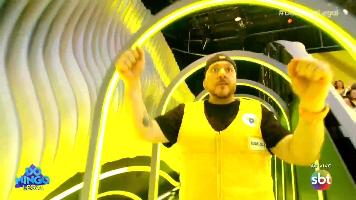 A man in a yellow vest with a name tag reading "RODRIGO" stands with arms raised inside a yellow neon-lit tunnel, with "DOMINGO LEGAL" and "SBT AO VIVO" logos visible.