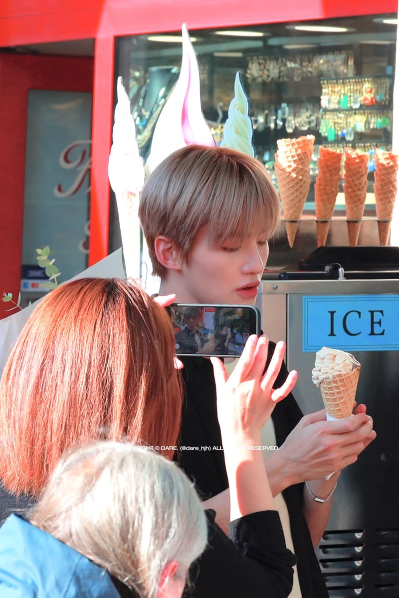 A young man with light brown hair holds a waffle cone of soft-serve ice cream while someone photographs him with a smartphone, standing in front of an ice cream stand with empty cones displayed and a watermark '© DARE. (@dare_hj) ALL RIGHTS RESERVED.'