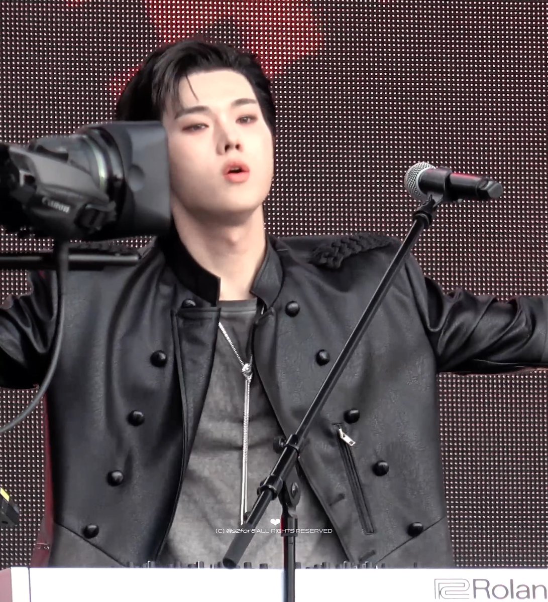 A performer in a black leather jacket stands at a microphone in front of a large camera on a tripod, with a watermark reading '(C) @s2for6ALL RIGHTS RESERVED' and 'Rolan' visible.