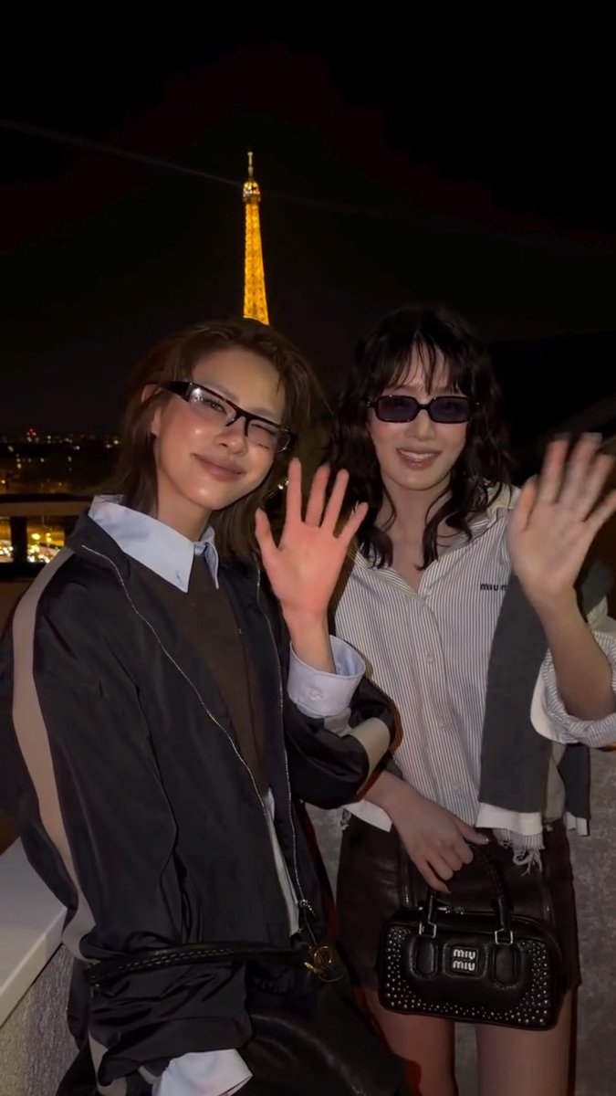 Two smiling women wearing glasses posing and waving in front of the illuminated Eiffel Tower at night, with one holding a black bag featuring "miu miu" logos.