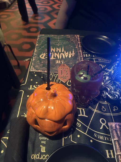 A Halloween-themed table setting with an orange jack-o'-lantern cup containing a straw, a skull glass, and a tablecloth featuring occult symbols, with a ticket numbered 13 visible.