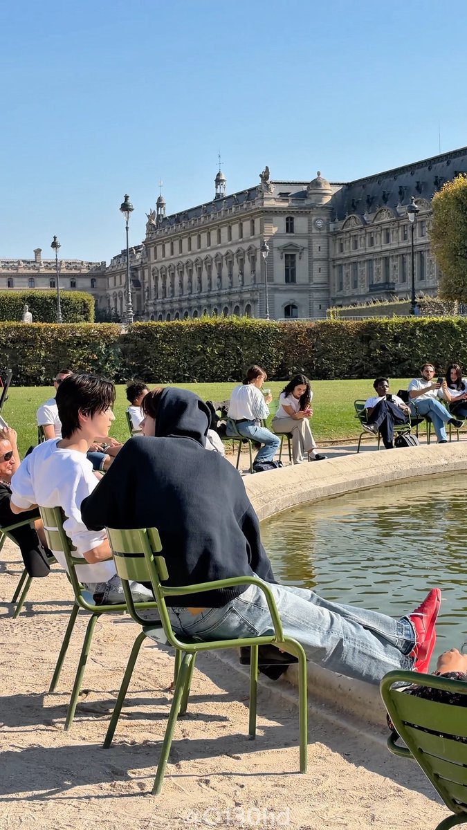 People sit on green metal chairs along the edge of a large reflecting pool in a park, with a grand historic European building in the background, possibly in Paris.
