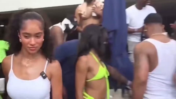 A young woman with dark hair pulled back, wearing a white tank top, stands at a crowded pool party while another woman in a neon green bikini stands nearby. The scene captures the social atmosphere referenced in the caption, with partygoers gathered around, though the woman highlighted in the post isn't immediately distinguishable in this frame.