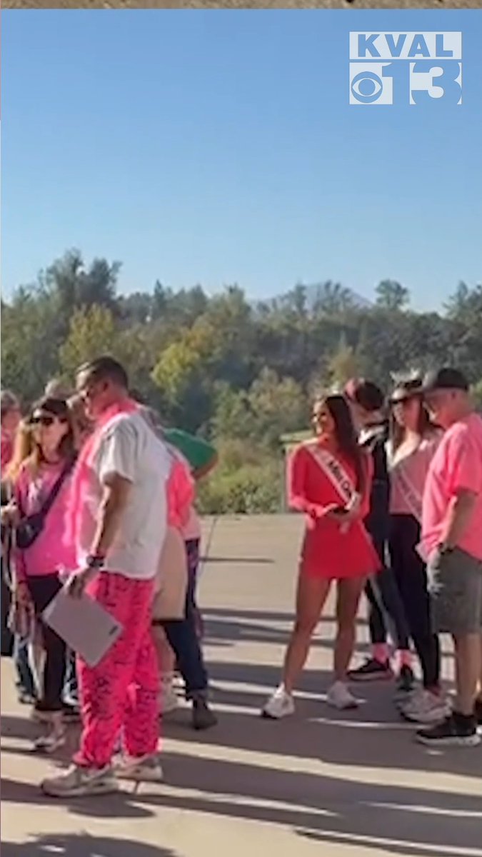 An outdoor gathering of people wearing pink shirts and accessories, including one person in a cowboy hat and another wearing a sash, with a 'KVAL CBS 13' news logo in the corner.
