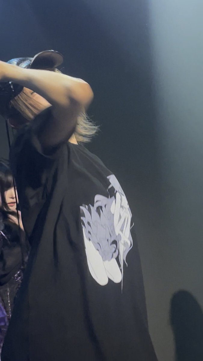 A person on stage wearing a black oversized t-shirt featuring a large white graphic design, with their arm raised near their face.