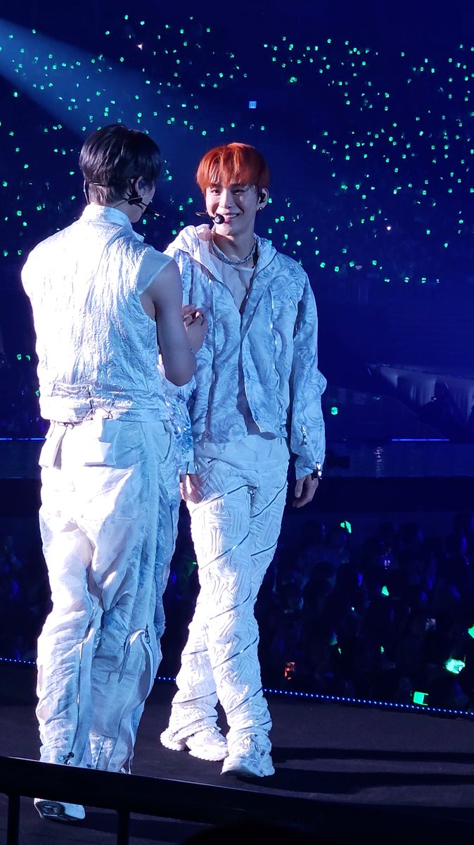 Two performers in matching white textured outfits on stage, with one smiling while facing the other, set against a backdrop of blue stage lighting and green light sticks.