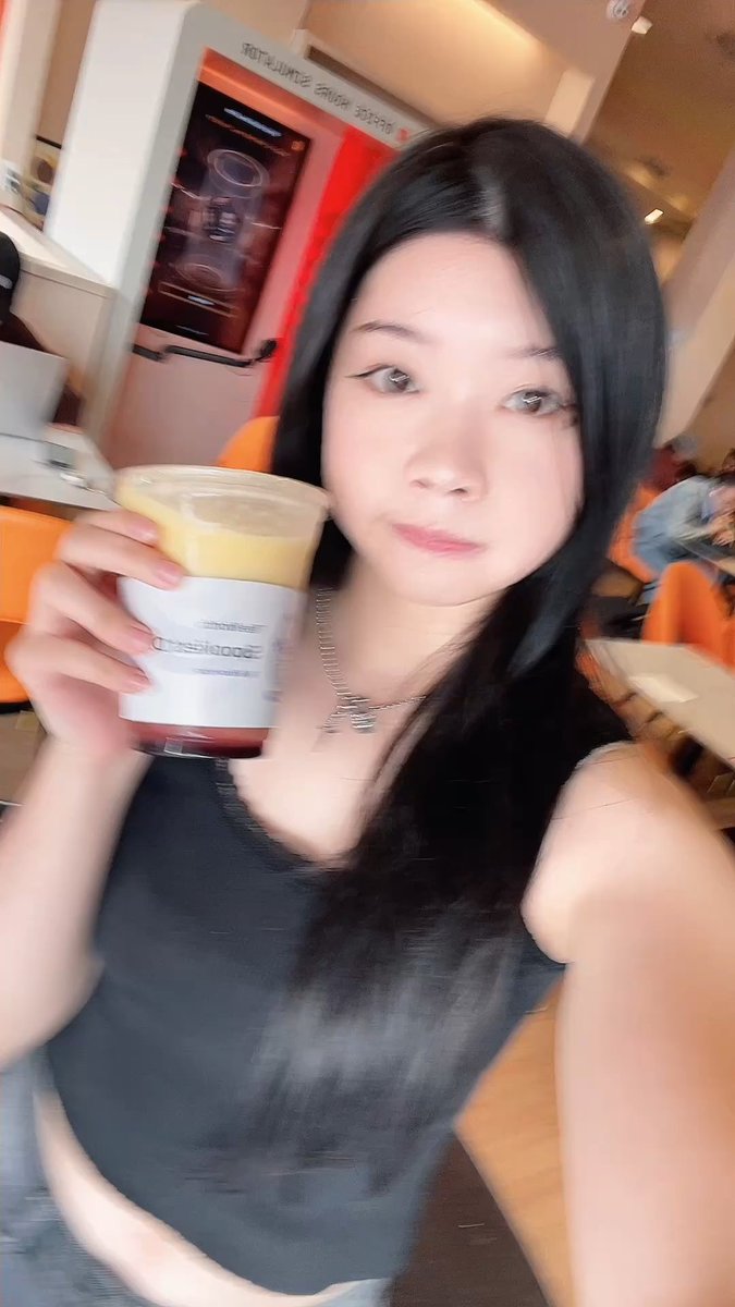 A young woman with long dark hair holds a beverage in a clear cup with a yellow topping, posing for a selfie indoors.