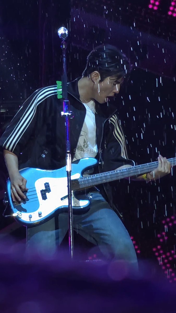 A musician intensely plays a blue and white bass guitar on stage in the rain, wearing a black jacket with white stripes over a graphic t-shirt.