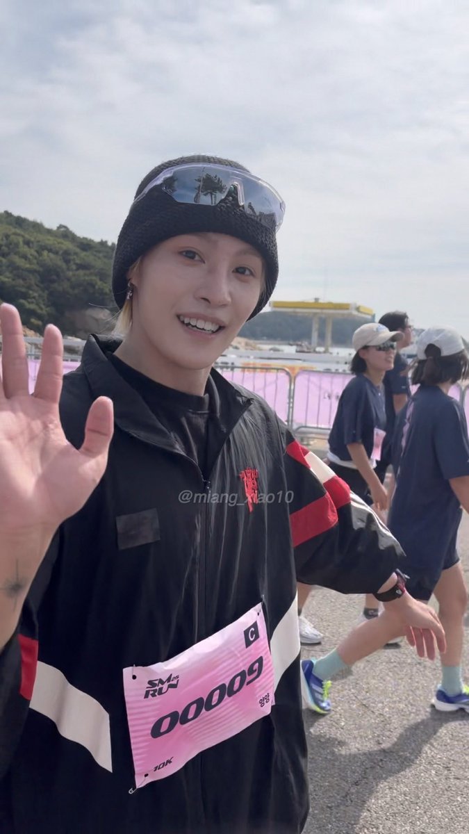 A smiling woman wearing a black beanie with sunglasses on top waves at the camera, sporting a black jacket and a race bib labeled 'SM2 RUN 00009' with '10K' and '2025' visible.