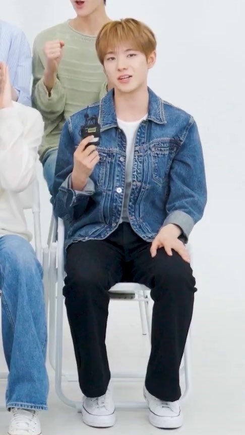 A young man in a denim jacket and black pants sits on a chair holding a small device, wearing white sneakers.