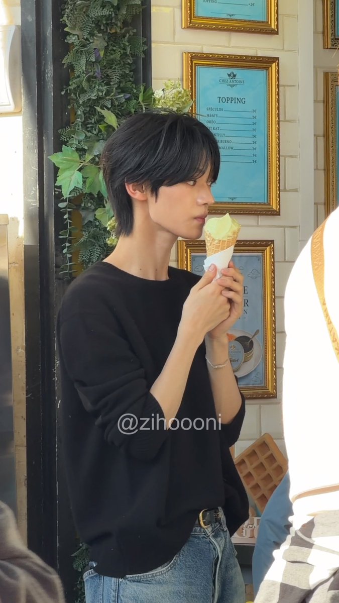 A young man with black hair wearing a black sweater eats a yellow ice cream cone in profile view inside a shop; a watermark '@zihooni' is visible on the image.