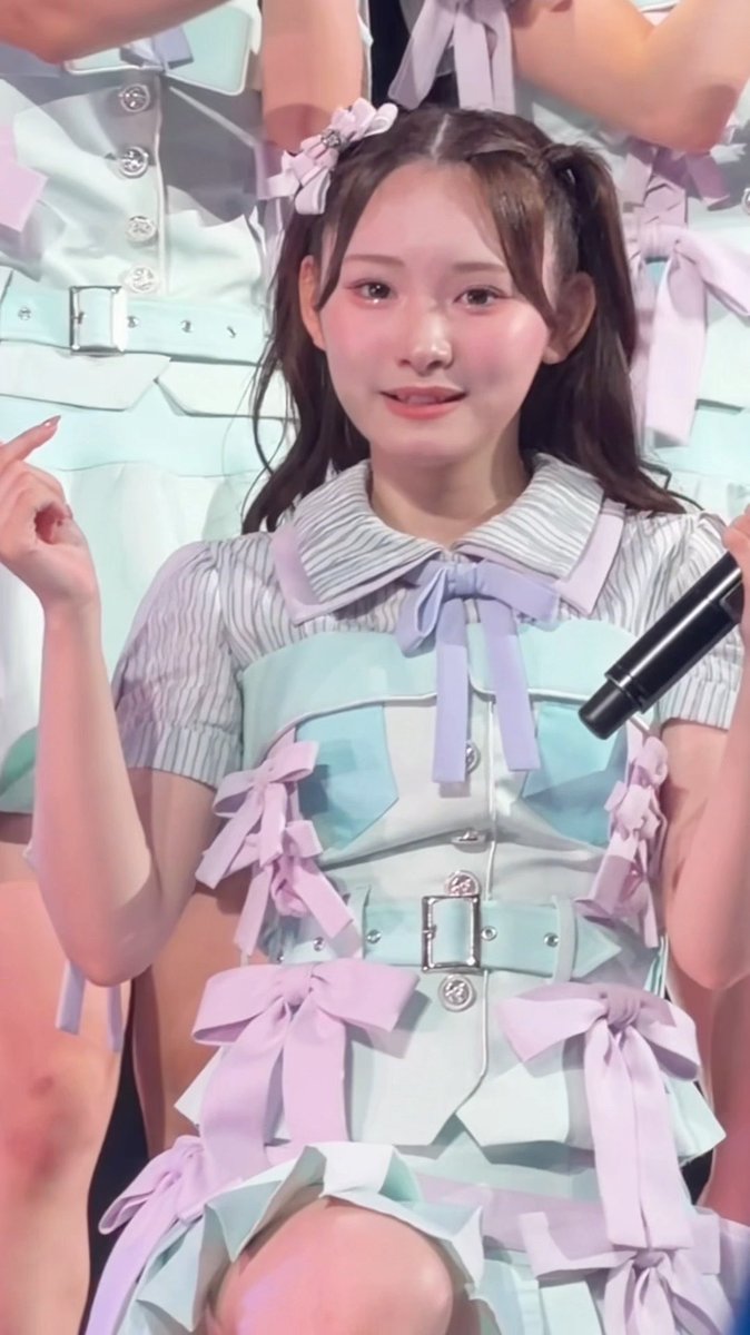 A young woman with pigtails wearing a pastel mint and lavender idol outfit with bow details smiles and gestures, holding a microphone.
