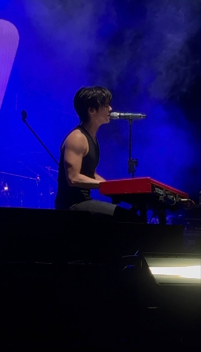 A man wearing glasses and a black tank top performing on stage, playing a red keyboard and singing into a microphone against a dramatic blue-lit background.