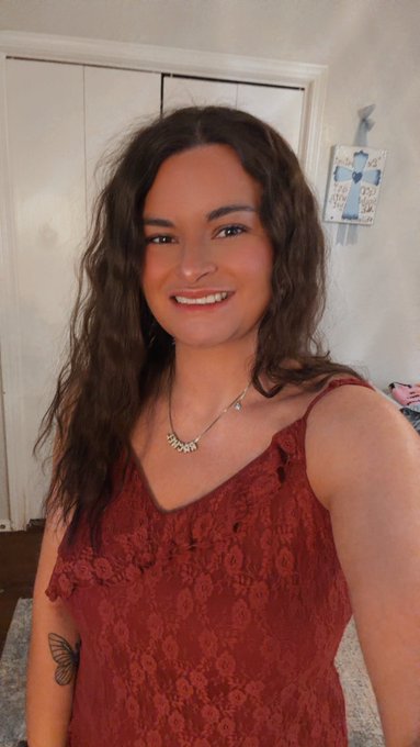 A woman with long wavy hair smiles at the camera while wearing a burgundy lace top and a silver name necklace, with a butterfly tattoo visible on her arm.