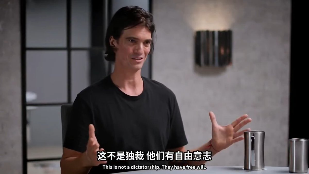 A man with dark shoulder-length hair wearing a black t-shirt sits in a modern room with gray walls, gesturing with both hands while speaking; Chinese and English subtitles read "这不是独裁 他们有自由意志 / This is not a dictatorship. They have free will."