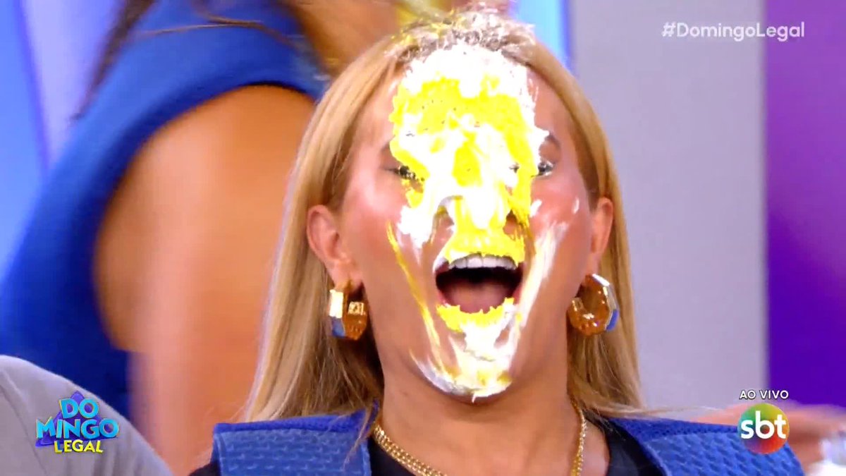 A blonde woman on a Brazilian TV show ('Domingo Legal' / 'sbt') reacts with an open mouth as a yellow and white substance is splattered across her face.