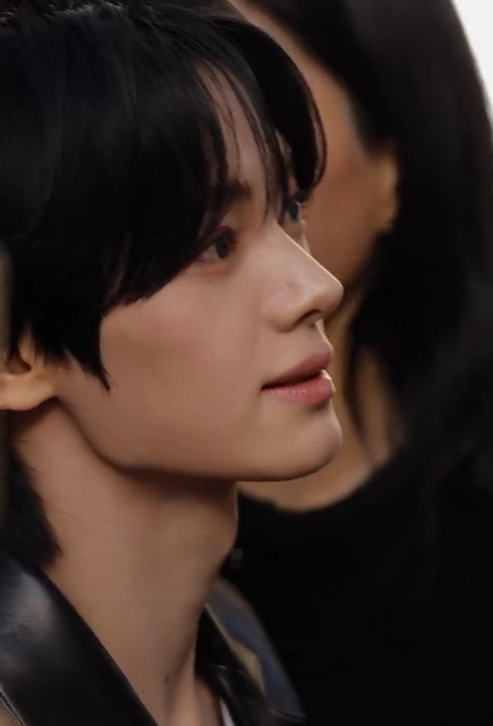 A close-up profile shot of a young man with dark hair looking off to the side, wearing a dark jacket.