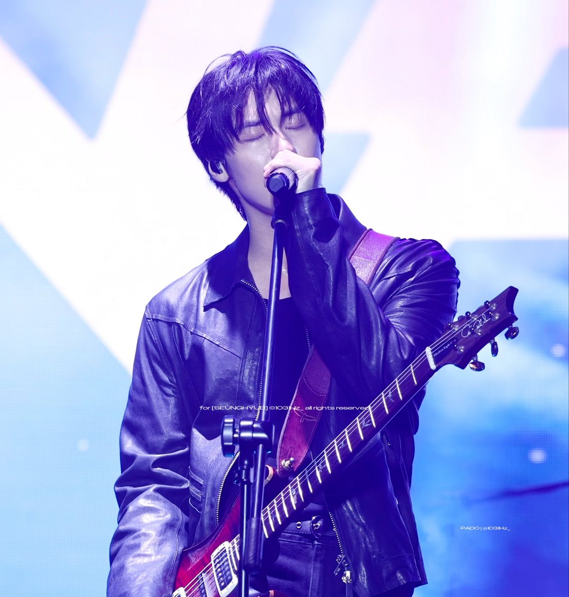 Concert photo of a male musician with dark hair wearing a black leather jacket, singing into a microphone while playing an electric guitar under blue stage lighting, watermarked 'for [SEUNGHYU] @103Hz all rights reserved'.