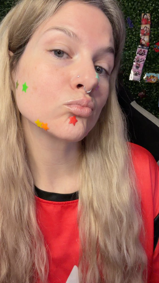 A close-up selfie of a young blonde woman wearing a red shirt, with colorful star-shaped face stickers (green, yellow, orange, red, purple) decorating her cheeks and chin.