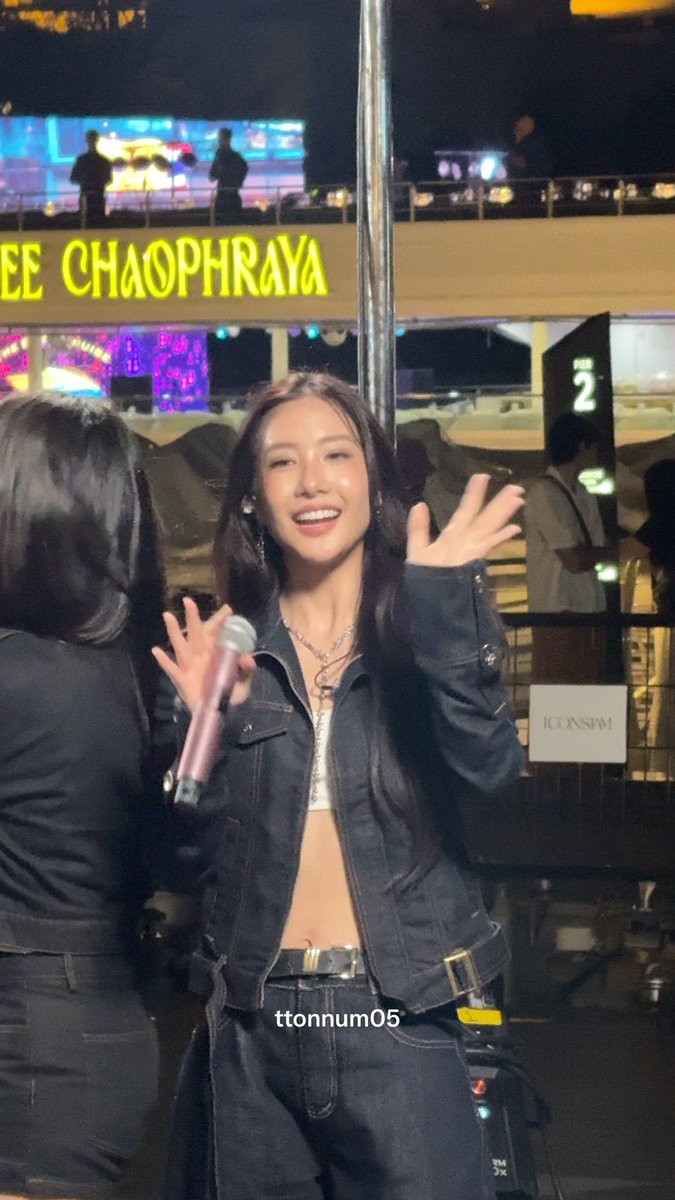 A smiling young woman with long dark hair holding a pink microphone and waving, with 'ICONSIAM' and 'CHAOPHRAYA' signage visible behind her; watermarked 'ttonnum05'.